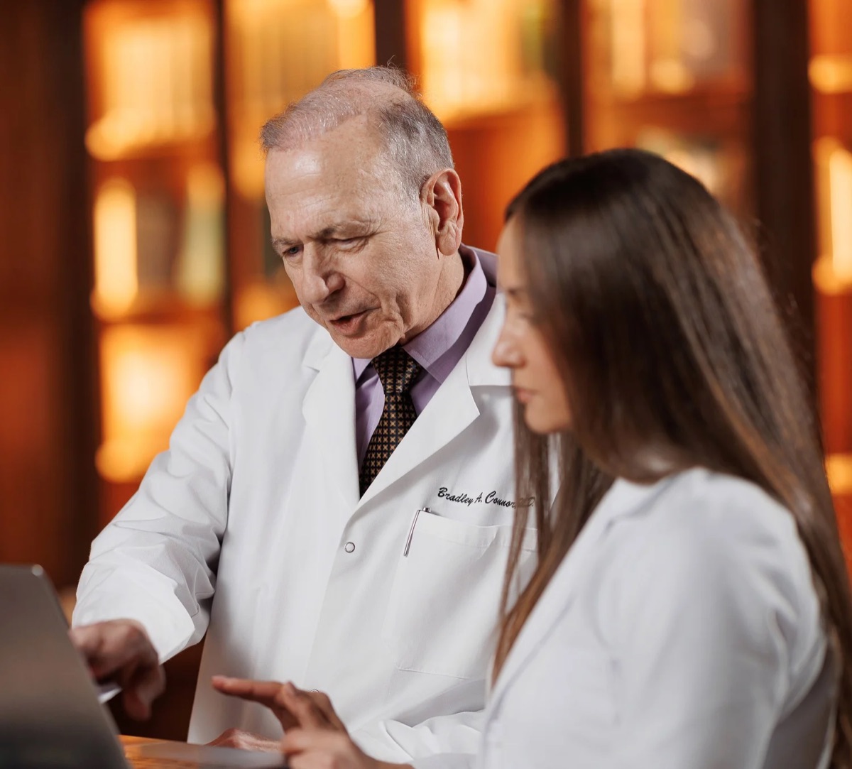 Dr. Bradley A. Connor and Marina Rogova, N.P., in white coats, looking at a laptop together in front of wooden bookshelves